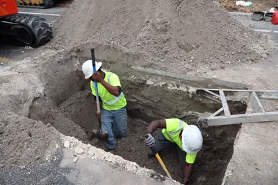 Centrel Services workers in full PPE hand-digging at the bottom of a deep, shielded trench to expose the existing sewer main.