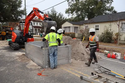 Kubota excavator preparing to lower an aluminum trench box into a deep excavation for sewer main repair.