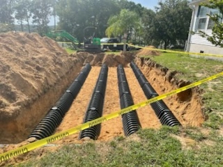 Wide trench with four rows of black plastic septic drain field chambers installed near a house with an excavator in the background