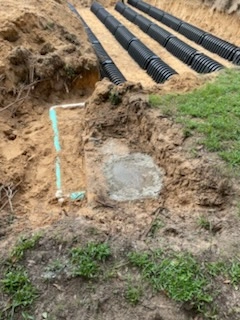 Partially buried concrete septic tank connected to a green PVC distribution pipe leading to black drain field chambers