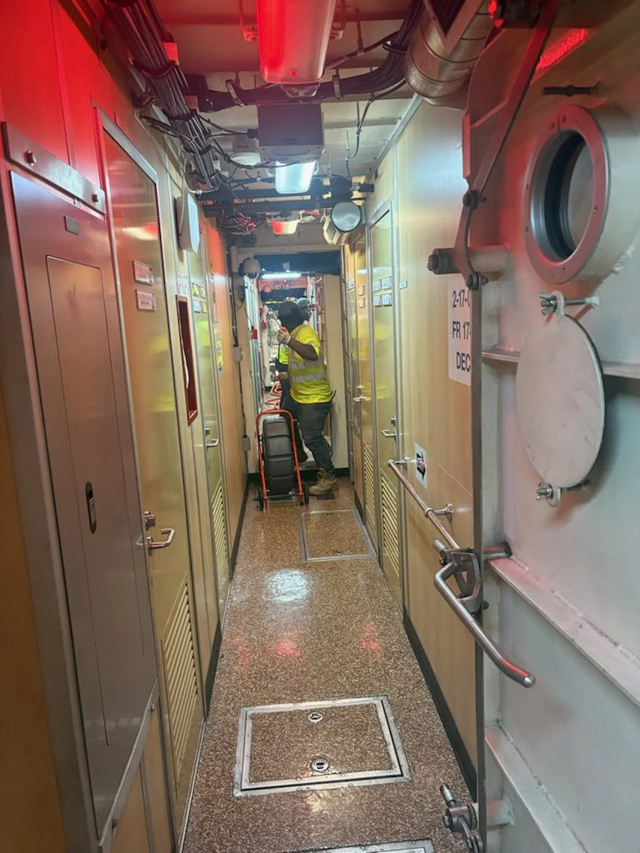 Narrow shipboard passageway with watertight doors and flush deck access plates while a technician moves equipment on a hand truck