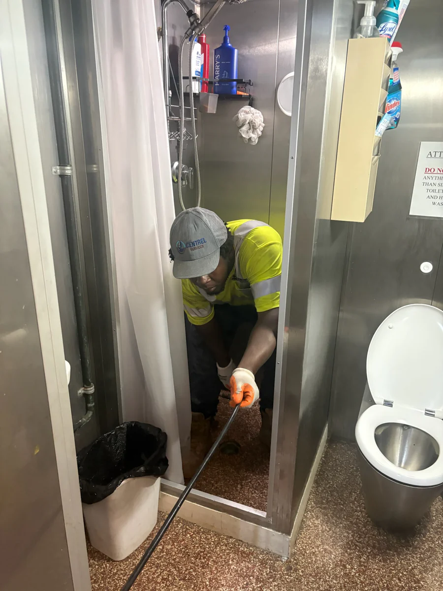 Centrel Services technician feeding a jetter hose into the drain of a stainless steel crew shower stall aboard a Coast Guard cutter
