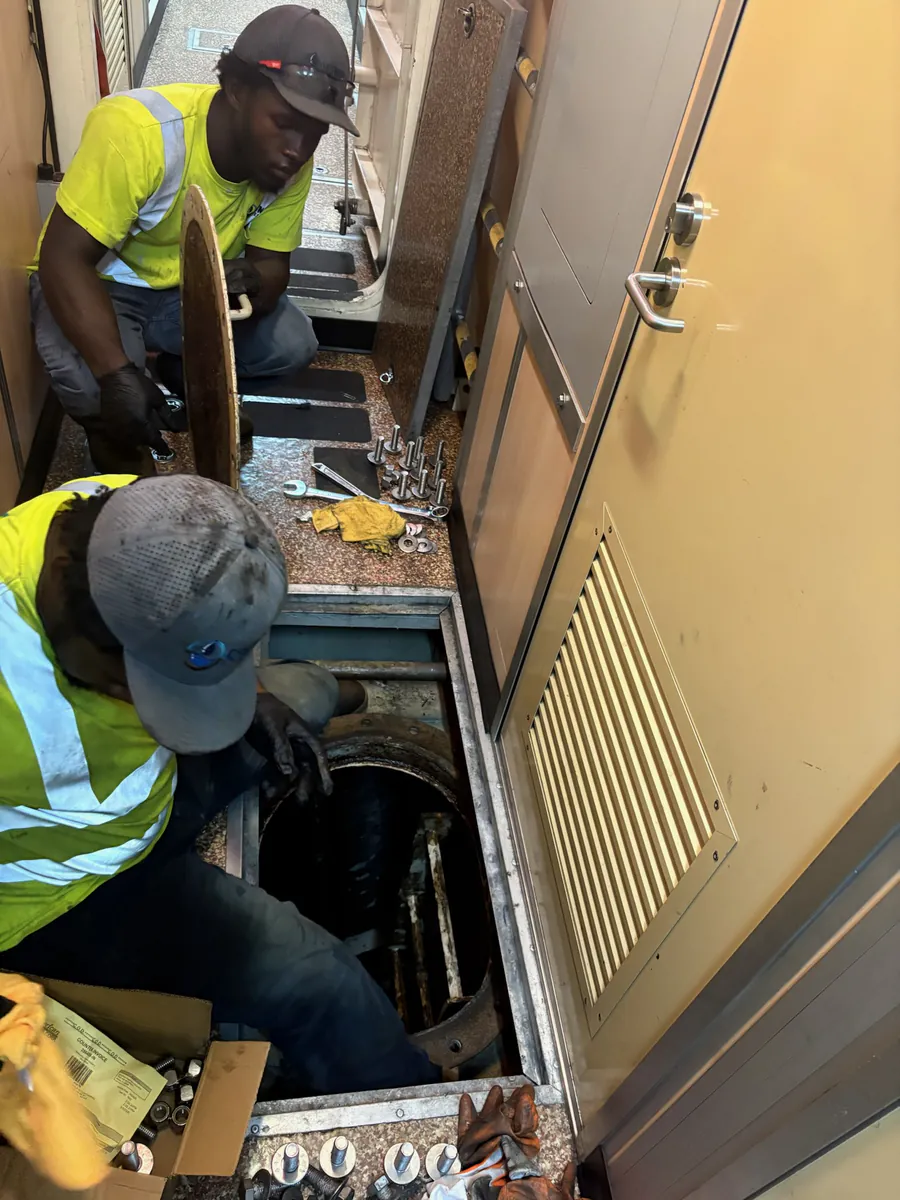 Two Centrel Services technicians in high visibility shirts lifting a bolted steel deck access plate to reach a grey water tank aboard a vessel