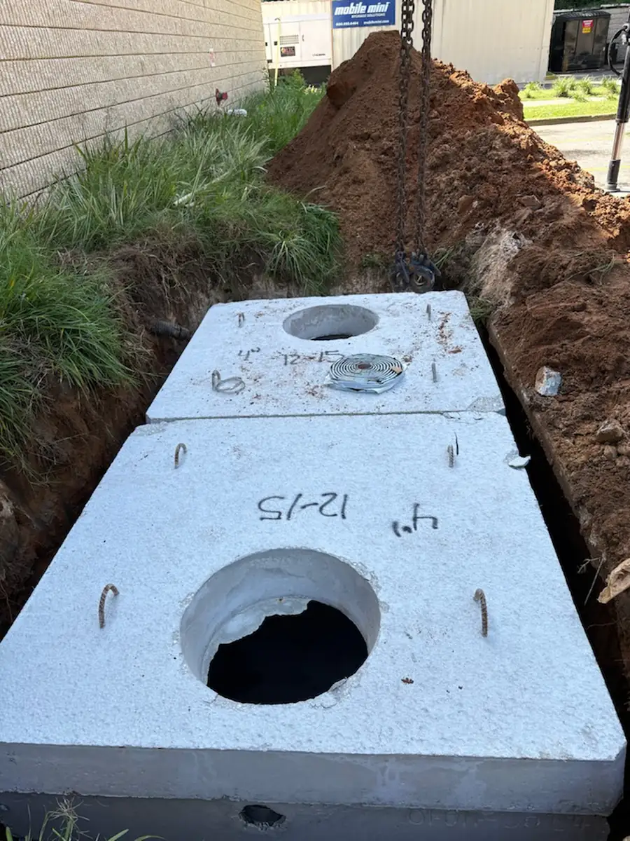 Precast concrete lids being lowered by chain onto a new grease interceptor with red clay spoil piled beside the trench