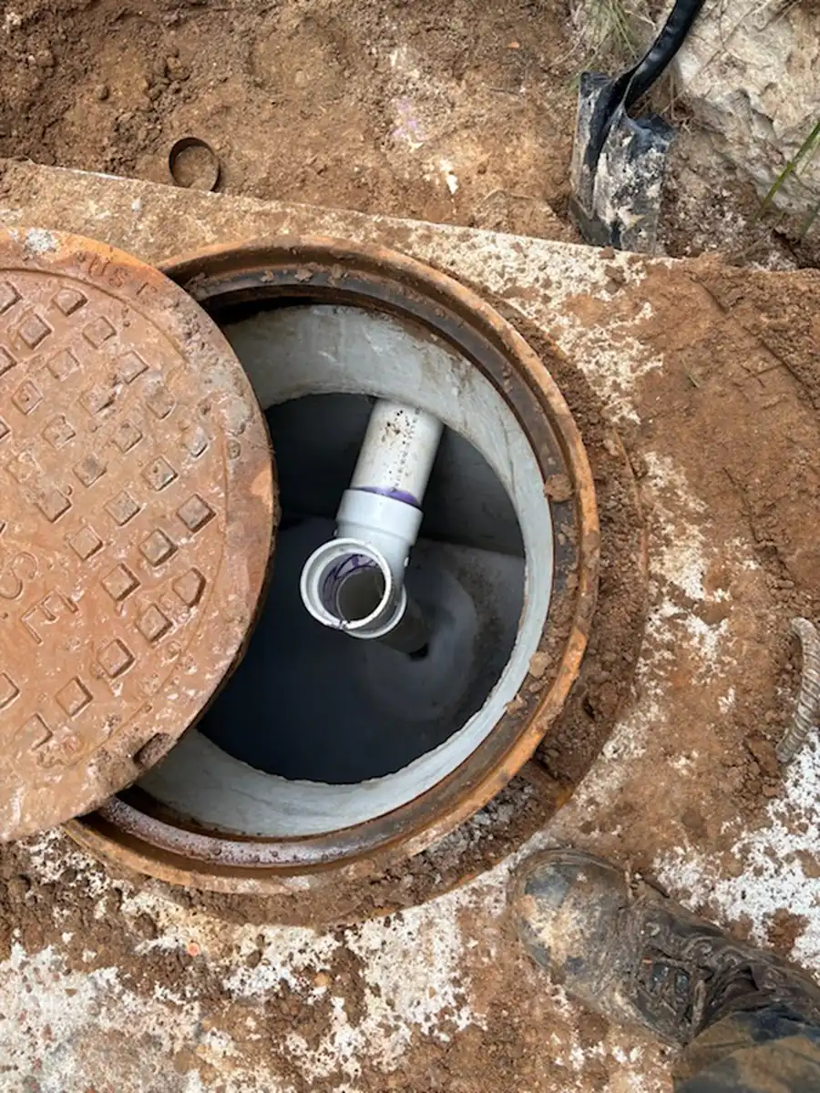 Cast iron access cover slid back from a finished grease trap riser showing the sanitary tee on the outlet below
