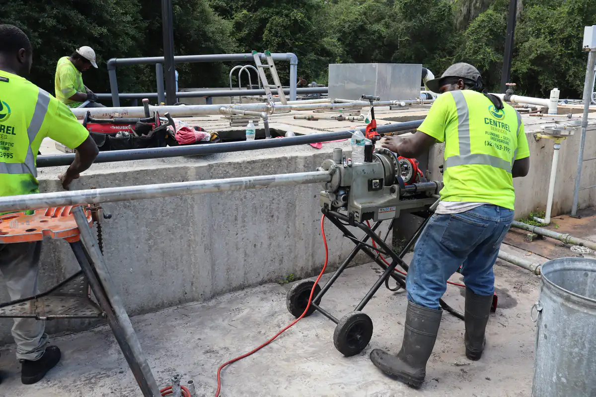 Centrel Services technician running galvanized pipe through a power threading machine while a coworker supports the pipe on a tripod stand