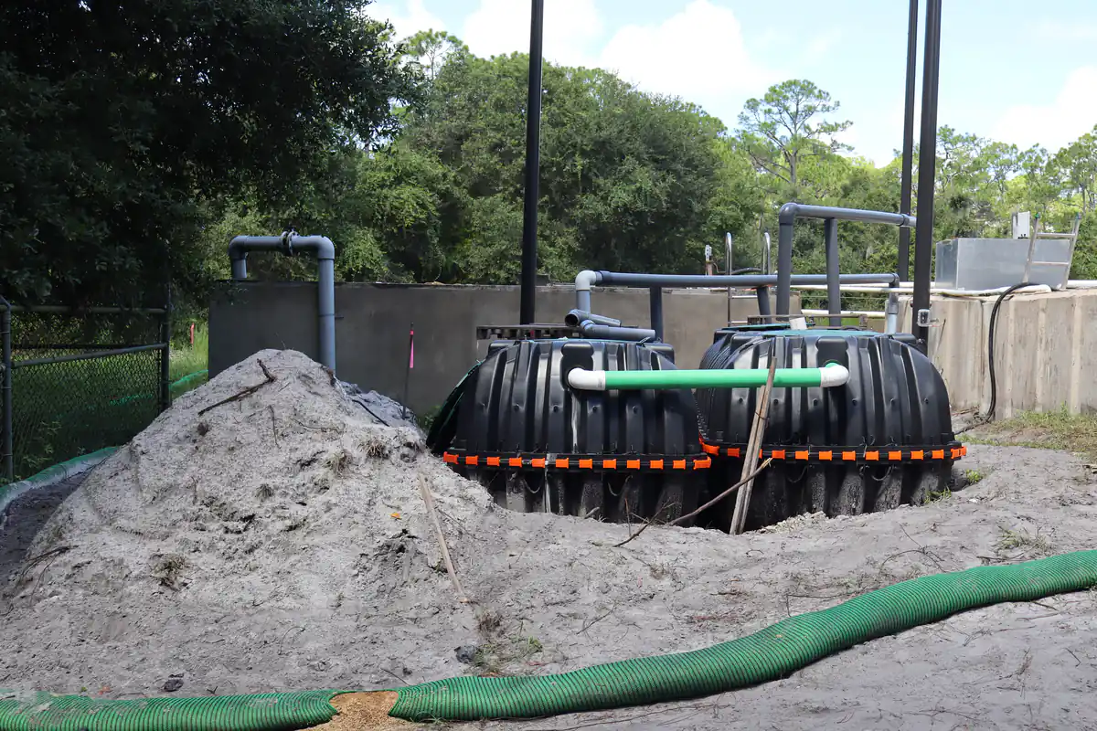 Two new black polyethylene septic tanks set side by side and joined by a green PVC crossover pipe beside a sand spoil pile