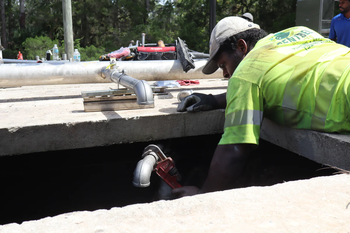 Technician leaning over a concrete basin wall to tighten a new galvanized elbow below the rim with a pipe wrench