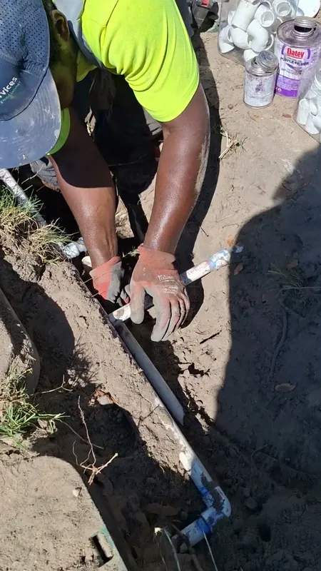 Technician in gloves connecting white PVC drain line at the bottom of a narrow trench